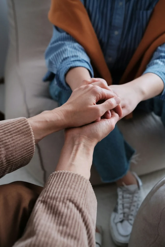Two people sit together holding hands, one offering comfort to the other, both wearing casual clothes and seated closely on a sofa.