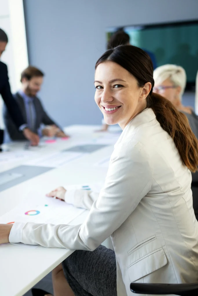 Woman in a white blazer smiles at the camera while seated at a meeting table with colleagues, papers and charts spread out in front of them.