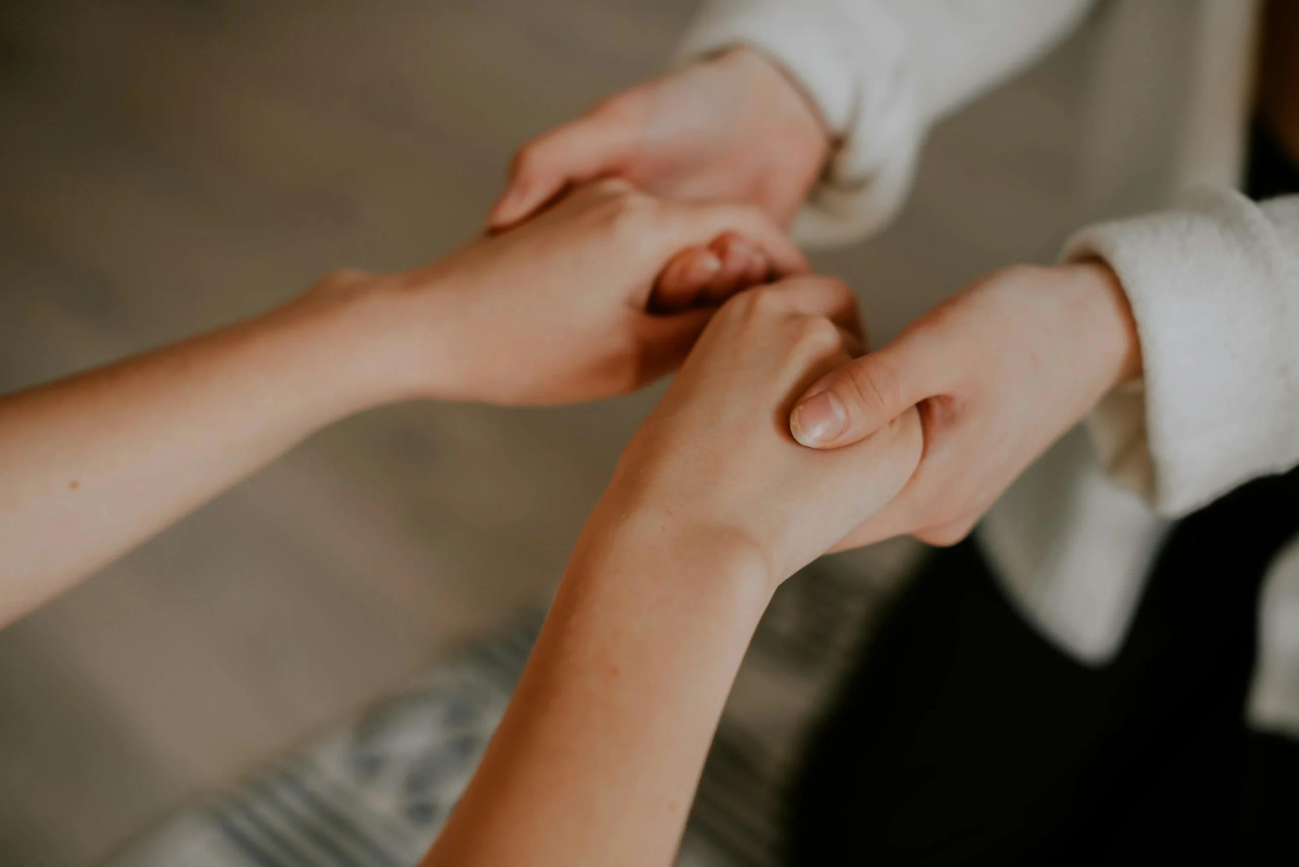 Two people are holding hands, with one pair of hands gently clasping the other. The background is neutral and out of focus.