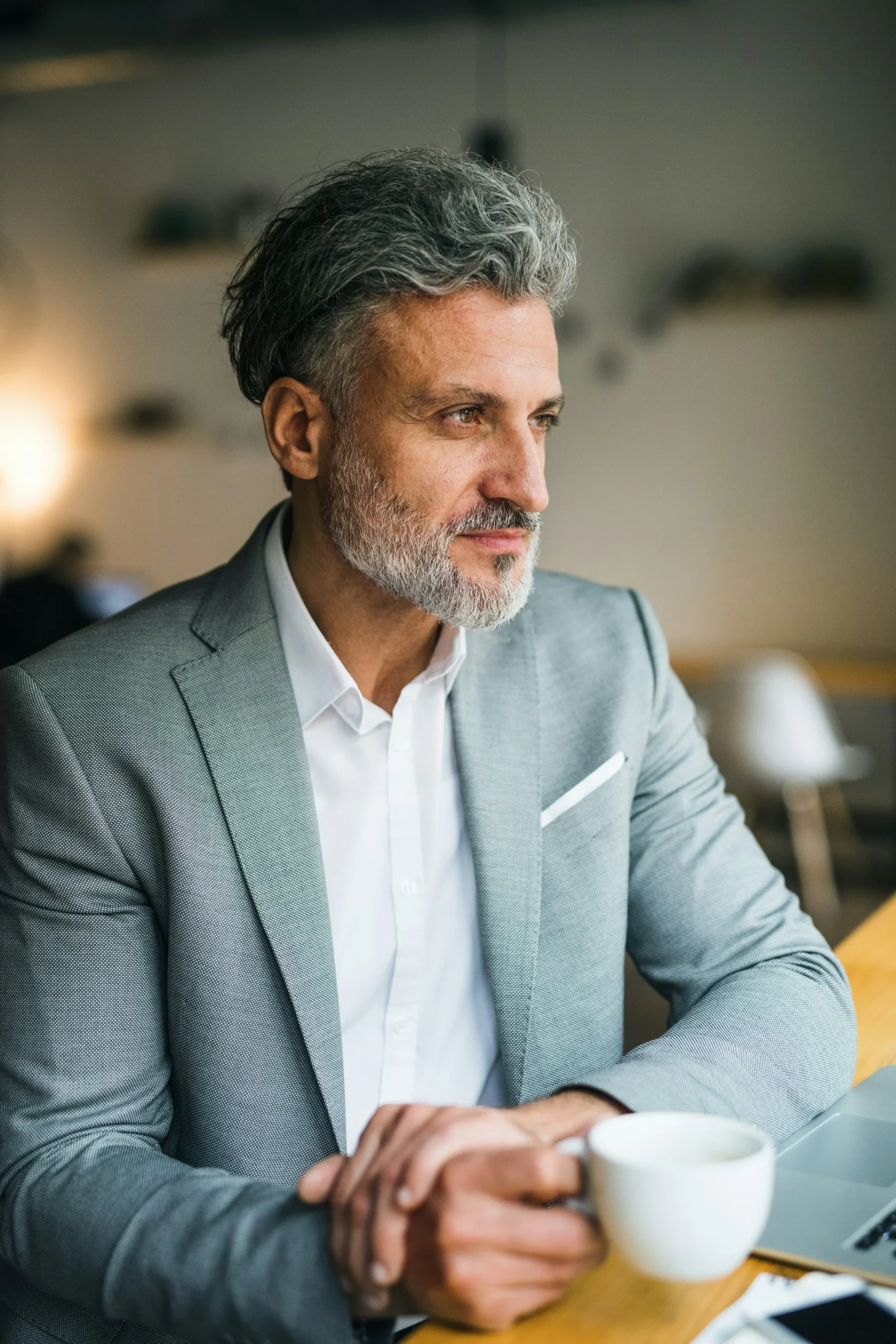 A man with grey hair and a beard, wearing a light grey suit and white shirt, sits at a table holding a white coffee cup, looking thoughtfully into the distance.