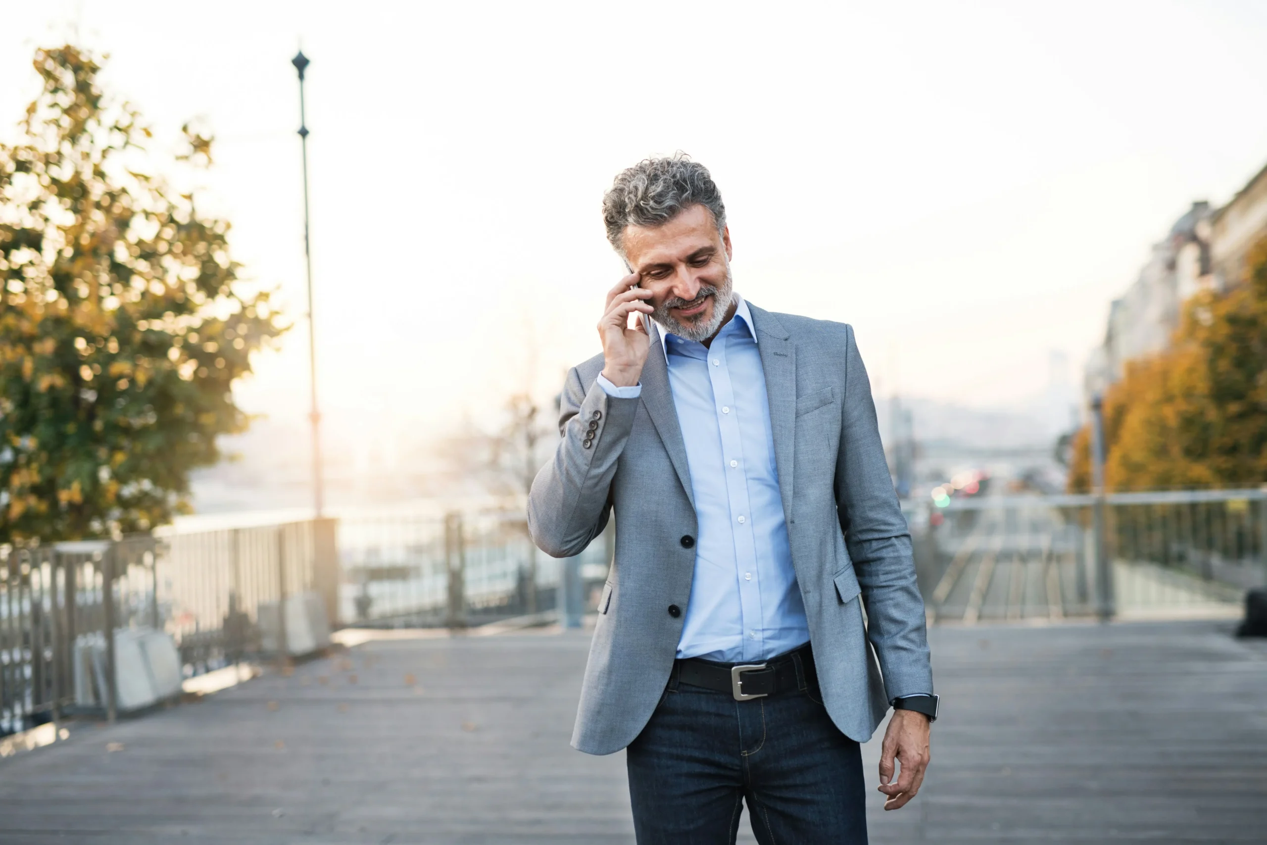 A man in a grey suit jacket talks on a mobile phone whilst walking outdoors on a boardwalk with trees and railings in the background.