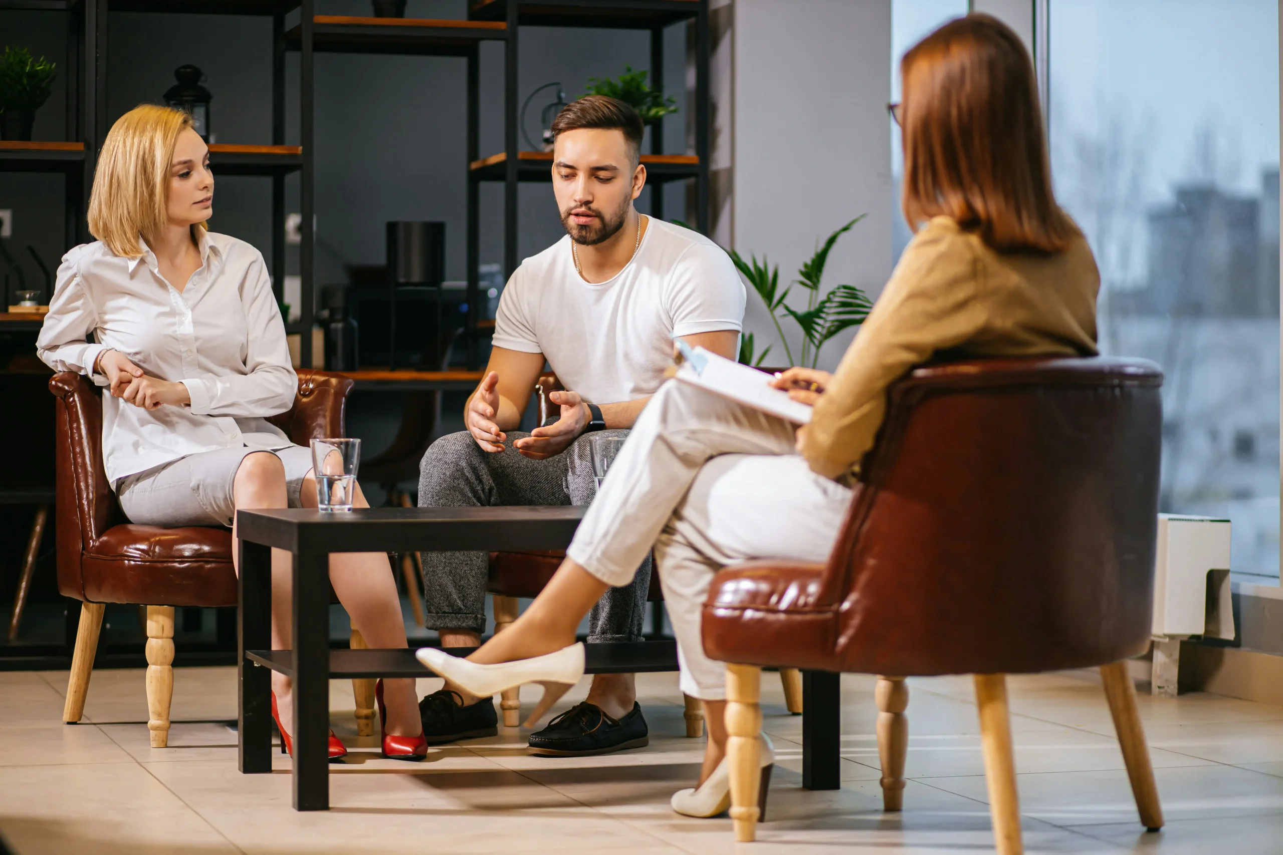 Three people sit in armchairs around a small table, engaged in a serious conversation; one person takes notes whilst the others talk and listen.
