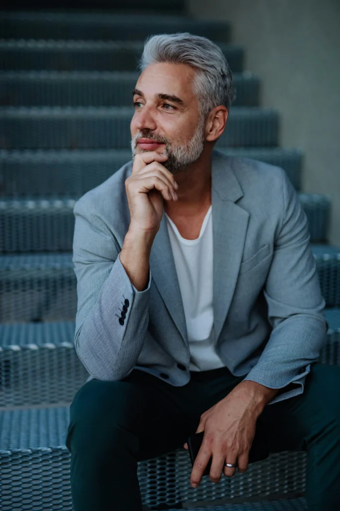 A man with grey hair and a beard, wearing a light grey blazer and white shirt, sits on metal steps with his hand resting on his chin, looking thoughtfully to the side.