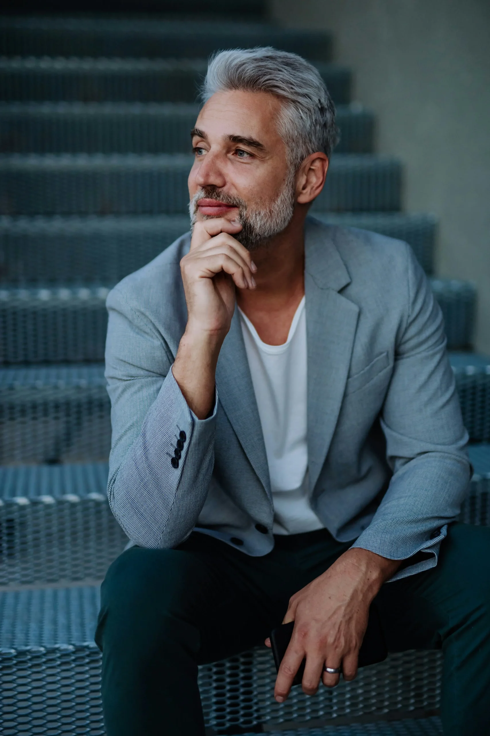 A man with grey hair and a beard, wearing a light grey blazer and white shirt, sits on metal steps with his hand resting on his chin, looking thoughtfully to the side.