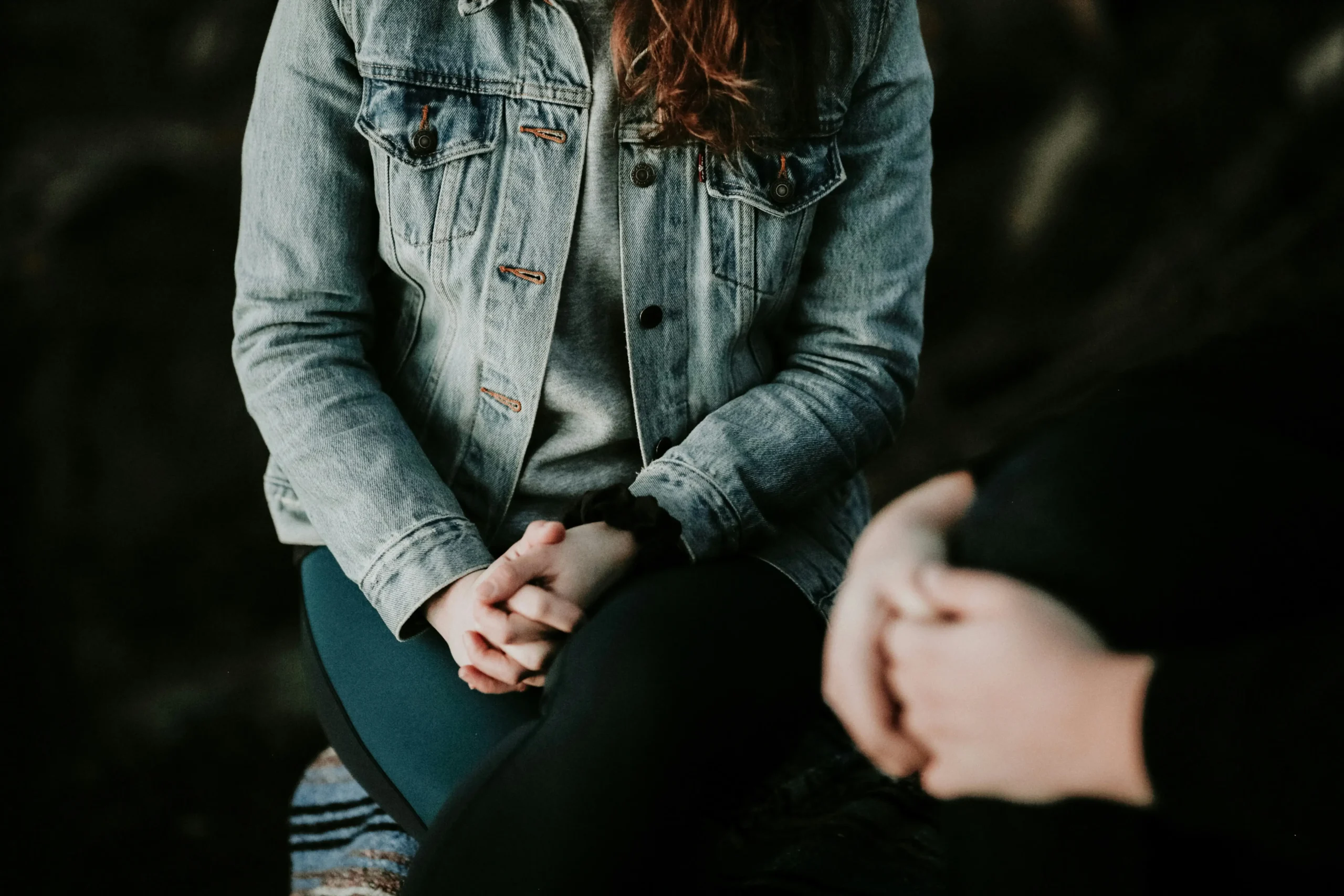 Two people sit close together, facing each other, with their hands clasped in their laps. One person wears a denim jacket, the other a dark long-sleeve top.