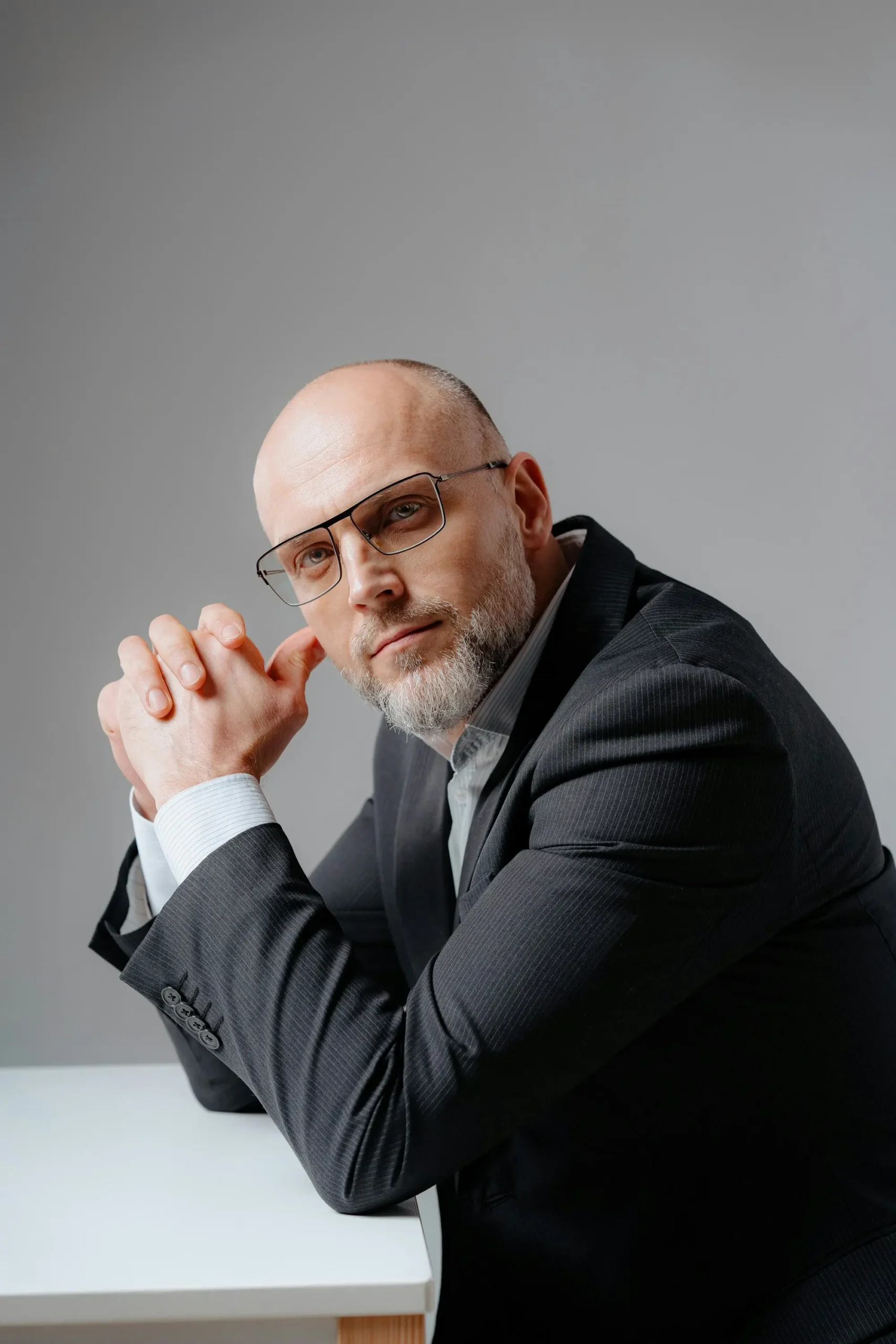 A middle-aged man with glasses, a bald head, and a trimmed beard sits at a table wearing a dark suit, looking at the camera with his hands clasped.