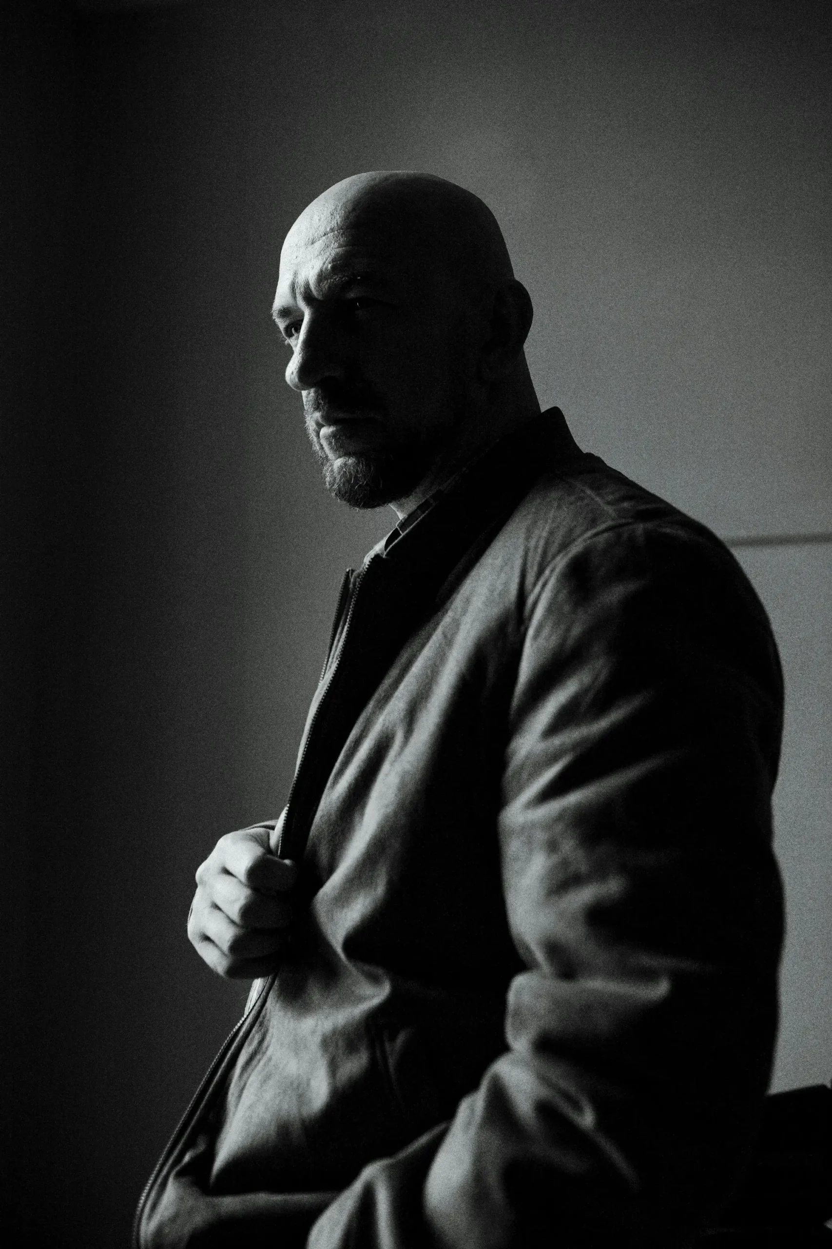 A bald man with a beard stands indoors, wearing a jacket and looking to the side in a dimly lit, shadowy environment.