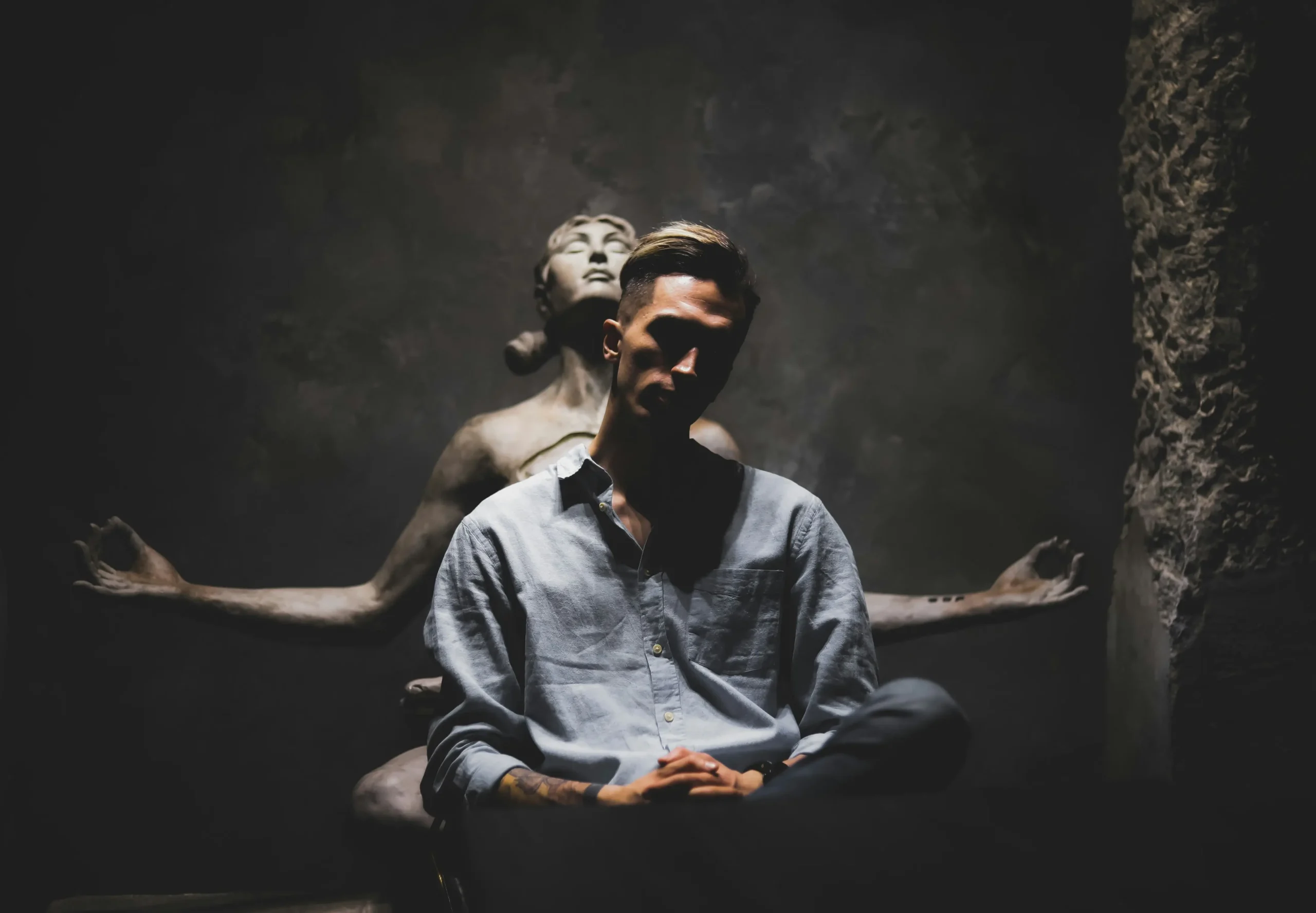 A man sits in dim lighting in front of a statue of a meditating figure, giving the illusion of extra arms behind him.